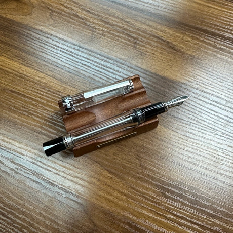 A clear TWSBI Vac700R fountain pen with a black cap and silver nib, resting on a wooden stand made of brown wood, placed on a wooden tabletop.