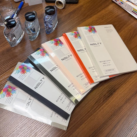 NOLTY A5 notebooks on a table in multiple colors including greige, black, khaki, pink, orange, and light gray.