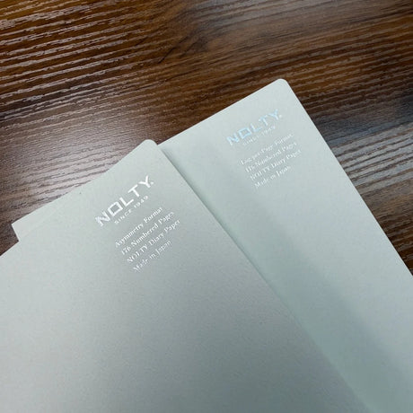 Closeup of two light gray NOLTY A5 notebook covers with text Asymmetry Format on one notebook and Log-per-Page on the second notebook, 176 Numbered Pages, NOLTY Diary Paper, Made in Japan.
