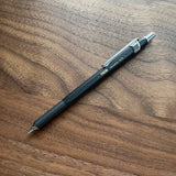 A black TWSBI 0.5 Precision Fixed Tip  mechanical pencil resting diagonally on a dark wood grain tabletop.