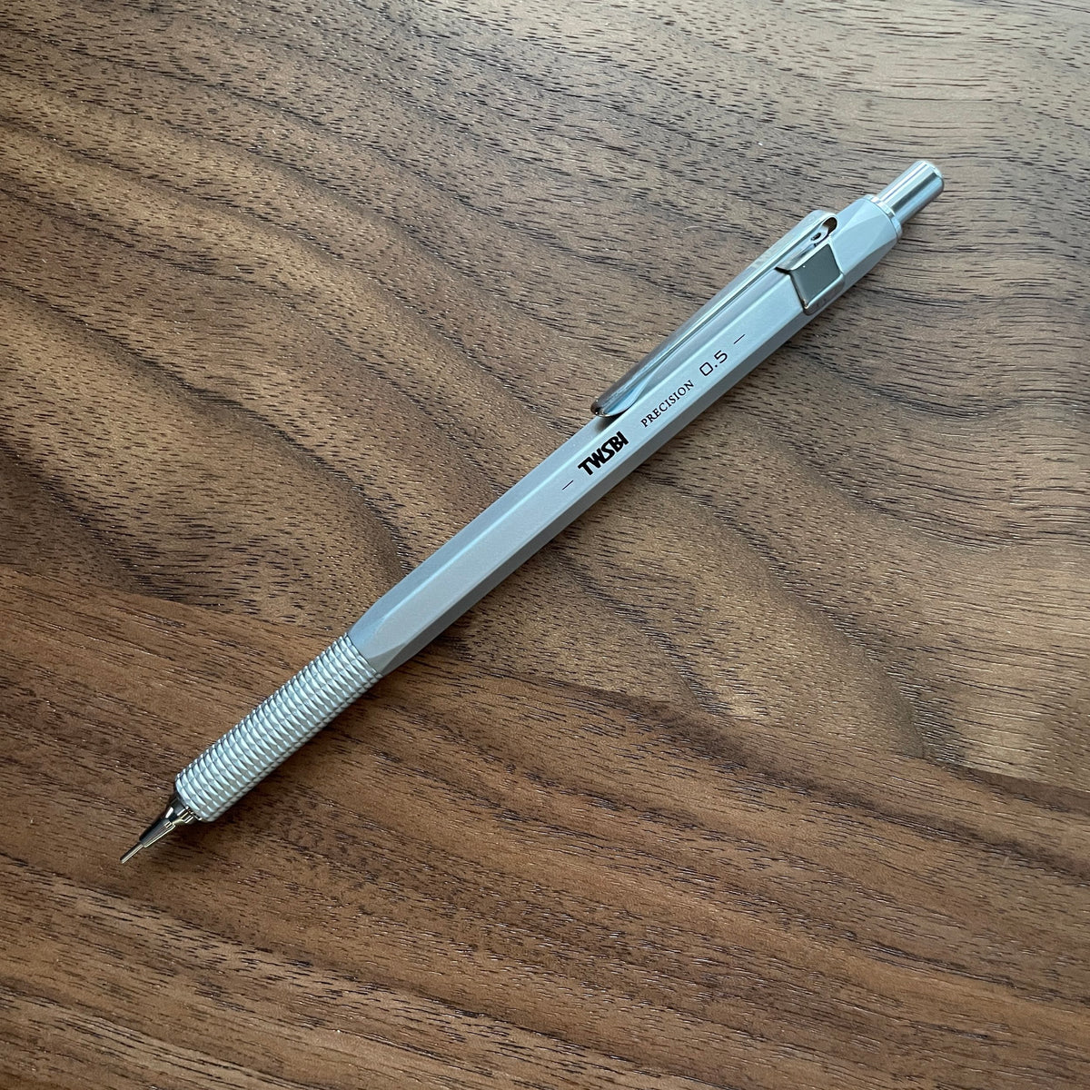 A silver TWSBI Precision Fixed Tip mechanical pencil with a textured grip and clip, resting diagonally on a brown wooden surface. 