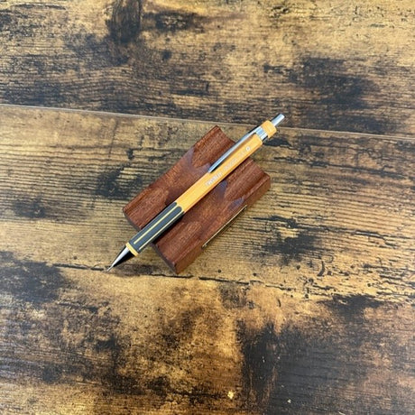 TWSBI Pagoda mechanical pencil with a yellow-orange barrel and silver metal accents, resting on a small wooden stand over a rustic dark wood tabletop.