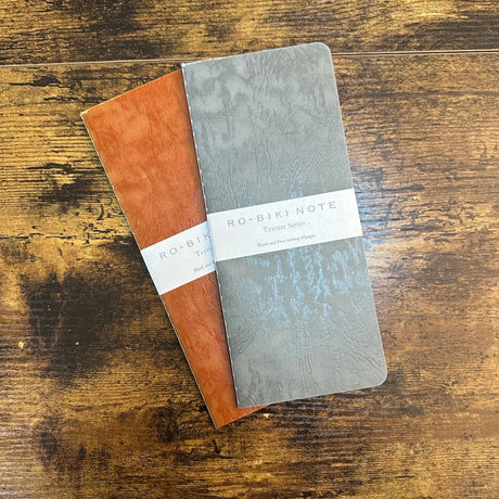 Two stacked standard Ro-Biki Note notebooks with textured covers in dark orange and gray and blank pages, A5 height and narrow width.