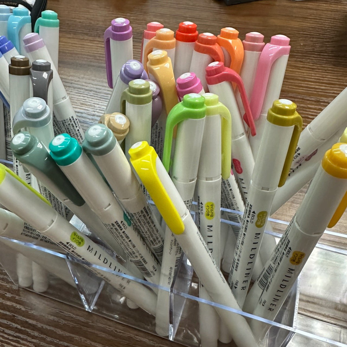 Several Zebra Mildliner highlighers in a variety of colors sitting in a clear acrylic pen holder on a table.