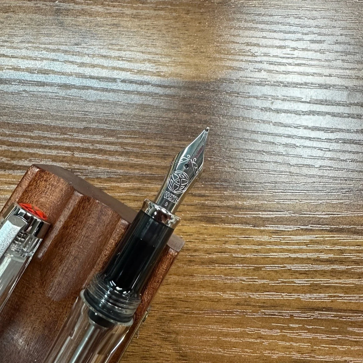 A close-up of a fountain pen nib and upper barrel resting on a wooden surface, with part of a wooden pen stand visible.
