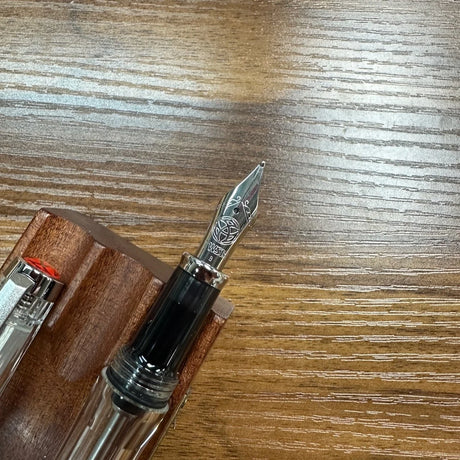 A close-up of a fountain pen nib and upper barrel resting on a wooden surface, with part of a wooden pen stand visible.
