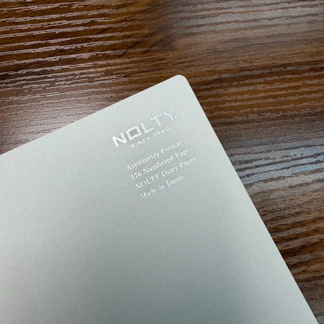 Closeup of light gray NOLTY A5 notebook cover with text Asymmetry Format, 176 Numbered Pages, NOLTY Diary Paper, Made in Japan.