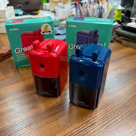 Two Carl Desktop Plastic Pencil Sharpeners in Red and Blue Plastic, shown with packaging on a desktop