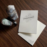 Letterpress greeting card with dark text 'Congratulations' and floral flourish design on thick, off-white paper with Skylab Letterpress logo and artwork credit on back.